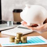 margem de contribuição - imagem de uma mão segurando um cofre em formato de porquinho sobre uma mesa com moedas empilhadas, papéis e uma calculadora