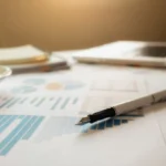 projeções financeiras - uma caneta em cima de papéis com dados financeiros sobre uma mesa com um notebook ao fundo